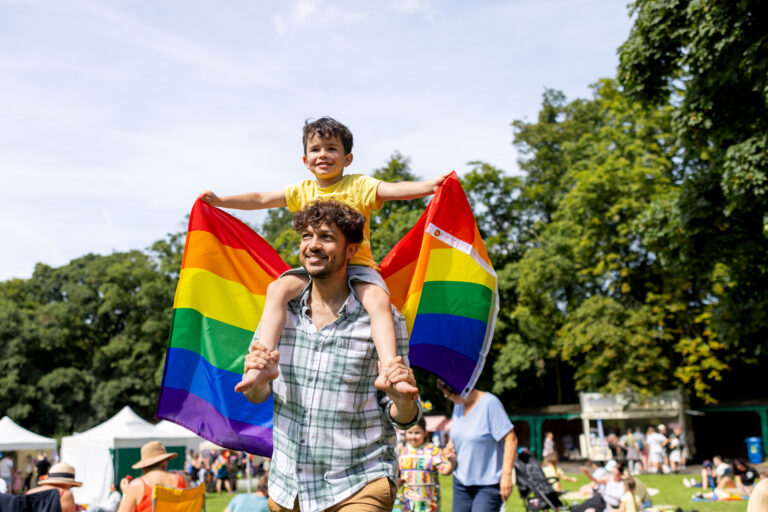 A waist-up shot of a joyful father and son spending quality time together in a sunny park. They have food vendors and the parade behind them. The setting is a bright, green outdoor space with trees and sunlight.